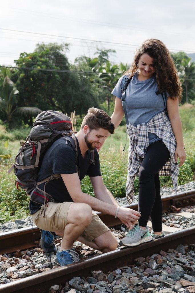 Positive man hiker squatting down and tying shoelaces of girlfriend standing on railroad placed in countryside during trip in sunny day